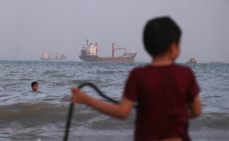 Children play in the water along the shore as a mix of bulk carriers, cargo ships, and service vessels sit offshore in the Strait of Hormuz off Bandar Abbas, Iran, Sunday.