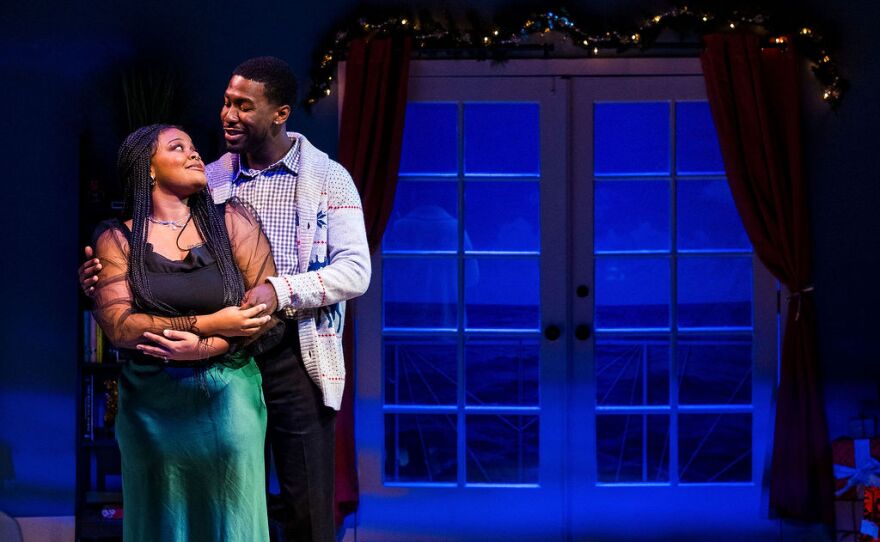 A woman and man embrace under a garland of holiday lights, near a set window at night, on stage. They have medium-brown to brown skin and dark hair, and are wearing festive clothes. The woman wears a satin green skirt and the man wears a Christmas sweater. They are gazing at each other.