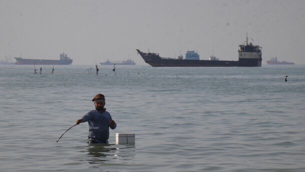 A man stands in the water, appearing to fish, as bulk carriers, cargo ships, and service vessels line the horizon in the Strait of Hormuz off Bandar Abbas, Iran, Monday, April 27, 2026.