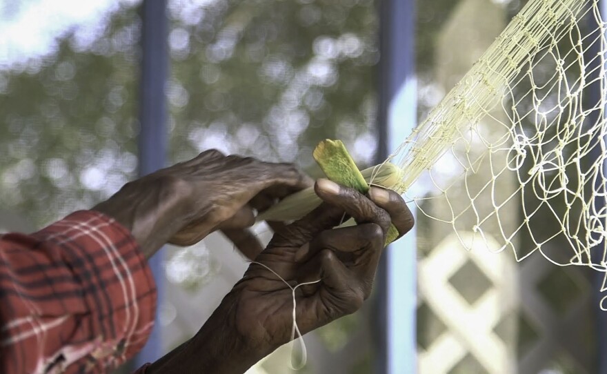 A medium shot of a pair of hands tying a net with a red, black, and white plaid sleeve visible. In the background, an out-of-focus trellis and trees are visible, with bright and sunny lighting.