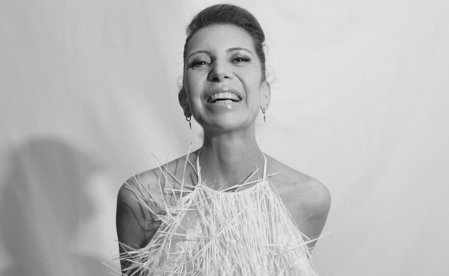 Musician Magos Herrera is shown in an undated photo. She is wearing a white dress with fringe-like strings blowing in the breeze. She is smiling wide. The photo is in black and white.