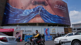 Vehicles drive past a billboard with an illustration showing the Strait of Hormuz and the sewn lips of President Trump in a square in downtown Tehran, Iran, May 2.