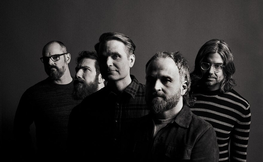 The five members of the band Death Cab for Cutie are shown in a black and white photo. Each has light skin, four have beards, two have glasses and one has long hair. The band members stand in a partial V shape and all but one are looking at the camera, with neutral expressions or smirking.