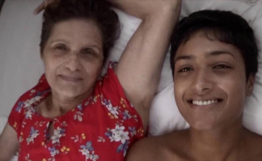 Chloe and Nana lay next to each other in bed, smiling and looking directly at the camera which looks back at them from above.