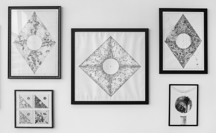 Five framed images are arranged on a wall. The art is in black and white, and is composed of hundreds of tiny lines, squiggles and characters formed into shapes, using negative and positive space to define borders. The largest, in the center, is diamond shaped, with a circle in the middle.