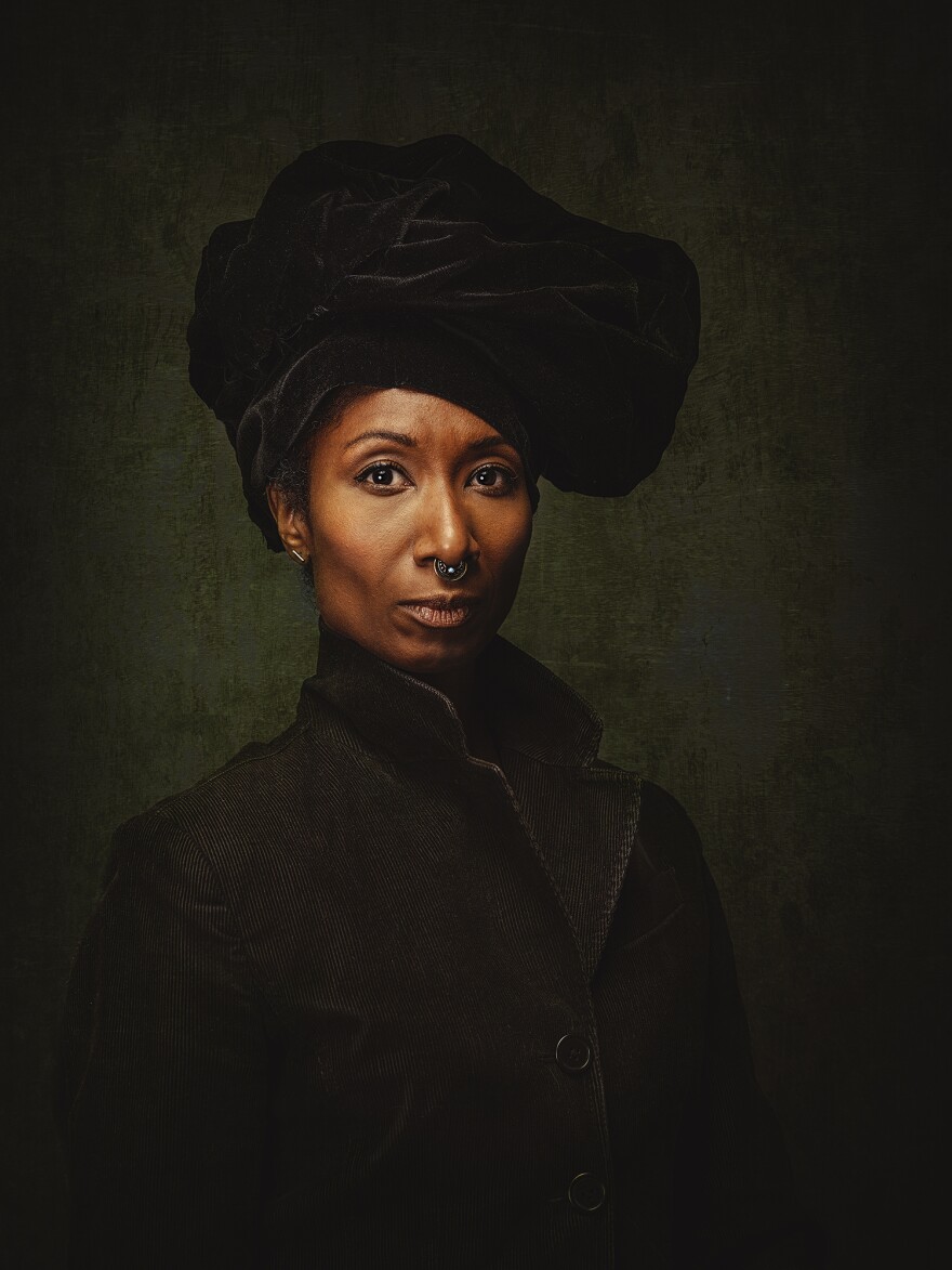 A woman wearing a black jacket, a black velvet hair-wrap style bonnet, and an ornamental nose ring is looking at the camera.