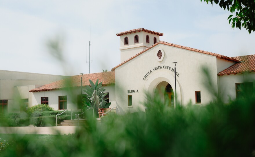 A white building with a red-tiled roof is seen through the blur of a green bush in the foreground.
