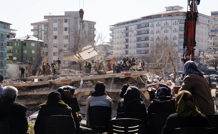 People watch as workers dig through a collapsed apartment building, looking for people who were inside the building when it collapsed in Islahiye, Turkey, on Sunday. In the week since a magnitude 7.8 earthquake and a series of powerful aftershocks hit Turkey and Syria, leveling cities like Islahiye, over 36,000 people have died.