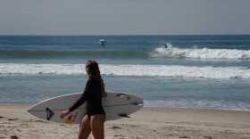 A woman carrying a surfboard walking on the beach in Oceanside as surfer catches a wave in the background, Sept. 19, 2025.