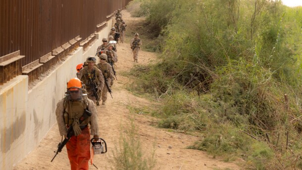 Marines with 1st Combat Engineer Battalion, armed with rifles and chainsaws, some dressed in blaze orange helmets and pants, walk in a column along the border fence move into position along the southern border to conduct vegetation clearance operations near Yuma, Ariz., Dec. 18, 2025.