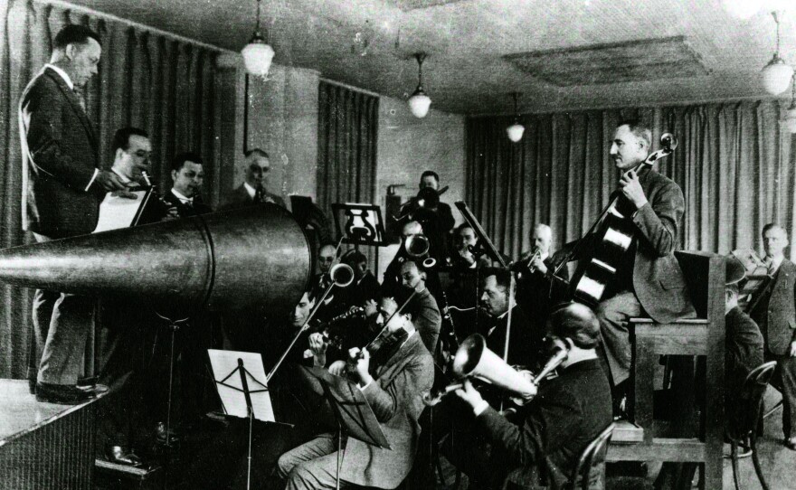 Before 1925, musicians like the Victor Orchestra, conducted by Rosario Bourdon, performed in front of a flared metal horn. An attached stylus would vibrate with the energy of the sound waves and etch them onto a wax rotating cylinder or disc — recording formats that are now very fragile.