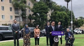 Mayor Todd Gloria discusses his plans to seek additional funding for the city's rental and utility assistance program. He is standing behind a podium thats reads, "A San Diego for All of Us." He is standing in front of a group of other people including, from left to right, Gilberto Vera, senior attorney, Legal Aid Society of San Diego, Nao Kabashima, executive director, Karen Organization of San Diego, City Council President pro Tem Stephen Whitburn,  District 3,  and City Councilmember Sean Elo-Rivera, District 9. They are standing on green grass. An apartment complex is visible in the background. 

