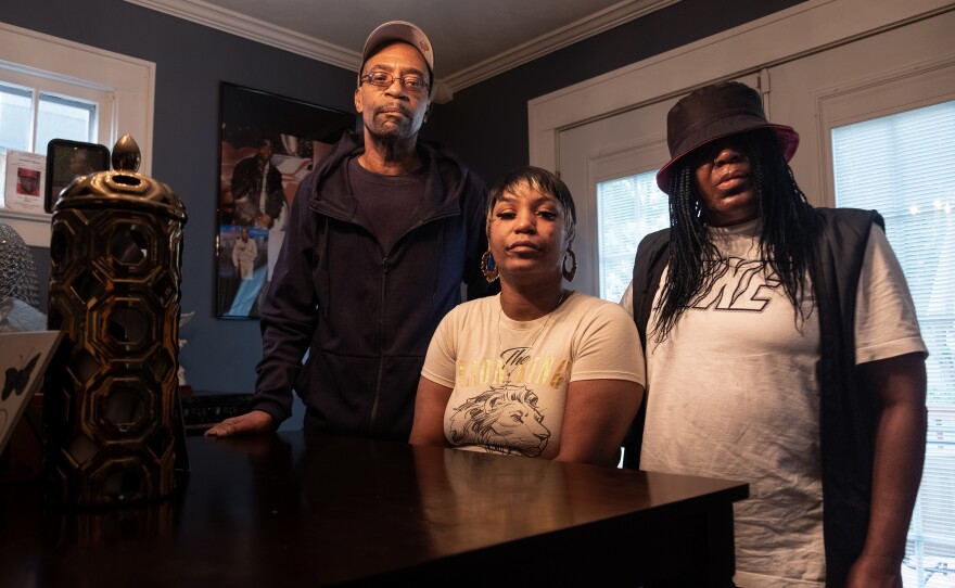 Ebony Everson (center), Bobby Everson's sister, stands with her father, who is also named Bobby Everson, and her mother Sabrina Everson. Photos of their son Bobby are on the wall behind them.