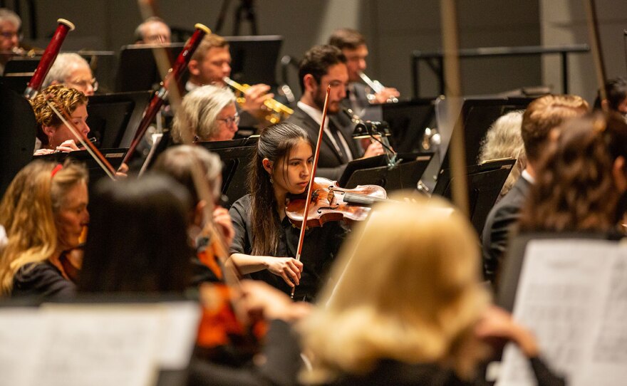 Performers from the La Jolla Symphony & Chorus are shown on stage in an undated photo. A violist with long brown hair is at the center of the photo, concentrating on the music and with the bow ready to strike the strings.