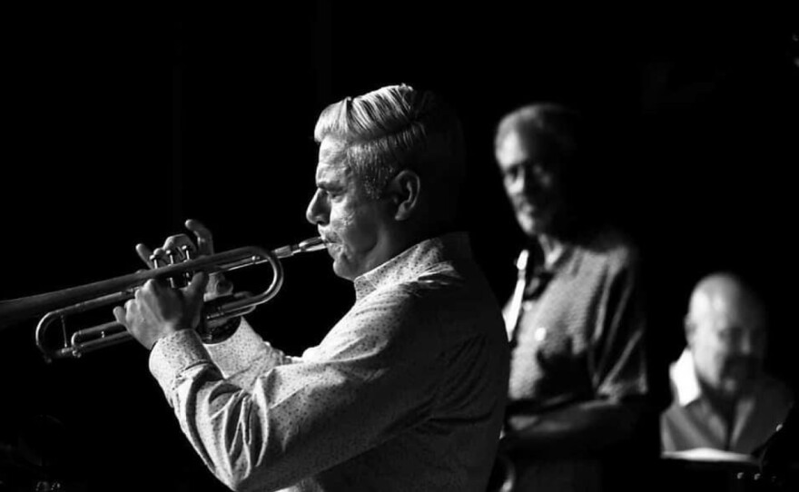 Gilbert Castellanos is shown in profile, playing trumpet, in a black and white photo with a black bacground. Out of focus in the background, other musicians look on.