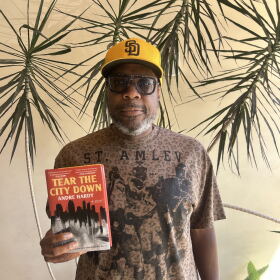 The image is a photograph of author and former NFL player Andre Hardy, as he holds up his novel "Tear the City Down" at KPBS on September 1, 2026.