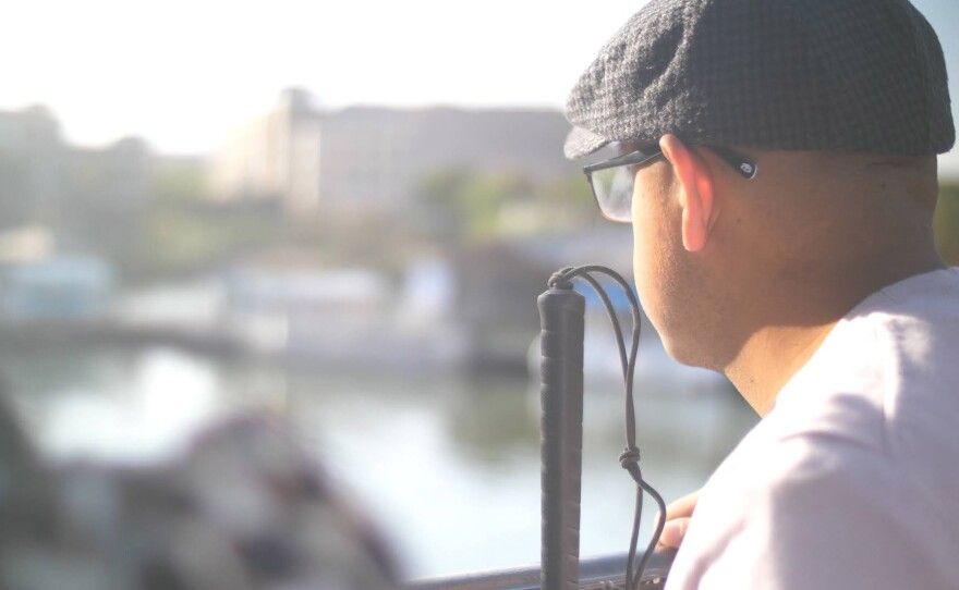 A man with glasses, gray beret, and light-colored shirt looks at an out-of-focus, daytime scenery. He has his back turned from the camera. Descripción: Un hombre con gafas, boina gris y camisa blanca observa un paisaje desenfocado. Está de espaldas a la cámara.